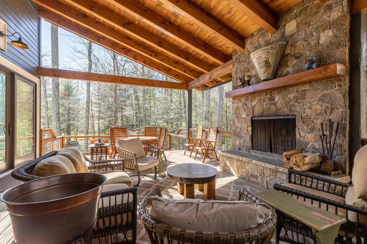 An exterior scene showcasing a cozy stone fireplace with wooden beams and cushioned seating, all set against an outdoor view filtered with natural light and forest glimpses. The mood is relaxed and inviting, perfect for enjoying cocktails or quiet evenings by the fire.