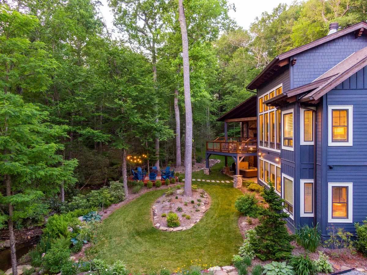 An exterior view of the home's blue facade and deck, with string lights overhead, a winding path through landscaped gardens, and hints of a fire pit. The scene is tranquil and cozy, inviting groups to gather outdoors, dine, or enjoy stargazing together.