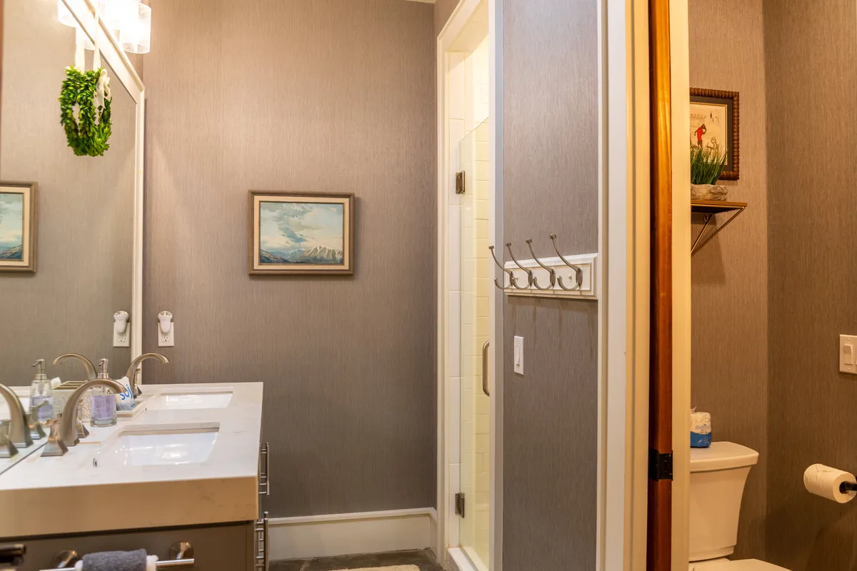 A serene bathroom scene with a double sink vanity, soft lighting, framed artwork, a spacious shower enclosure, and ample towel hooks. The layout supports both relaxation and practical use by multiple guests.