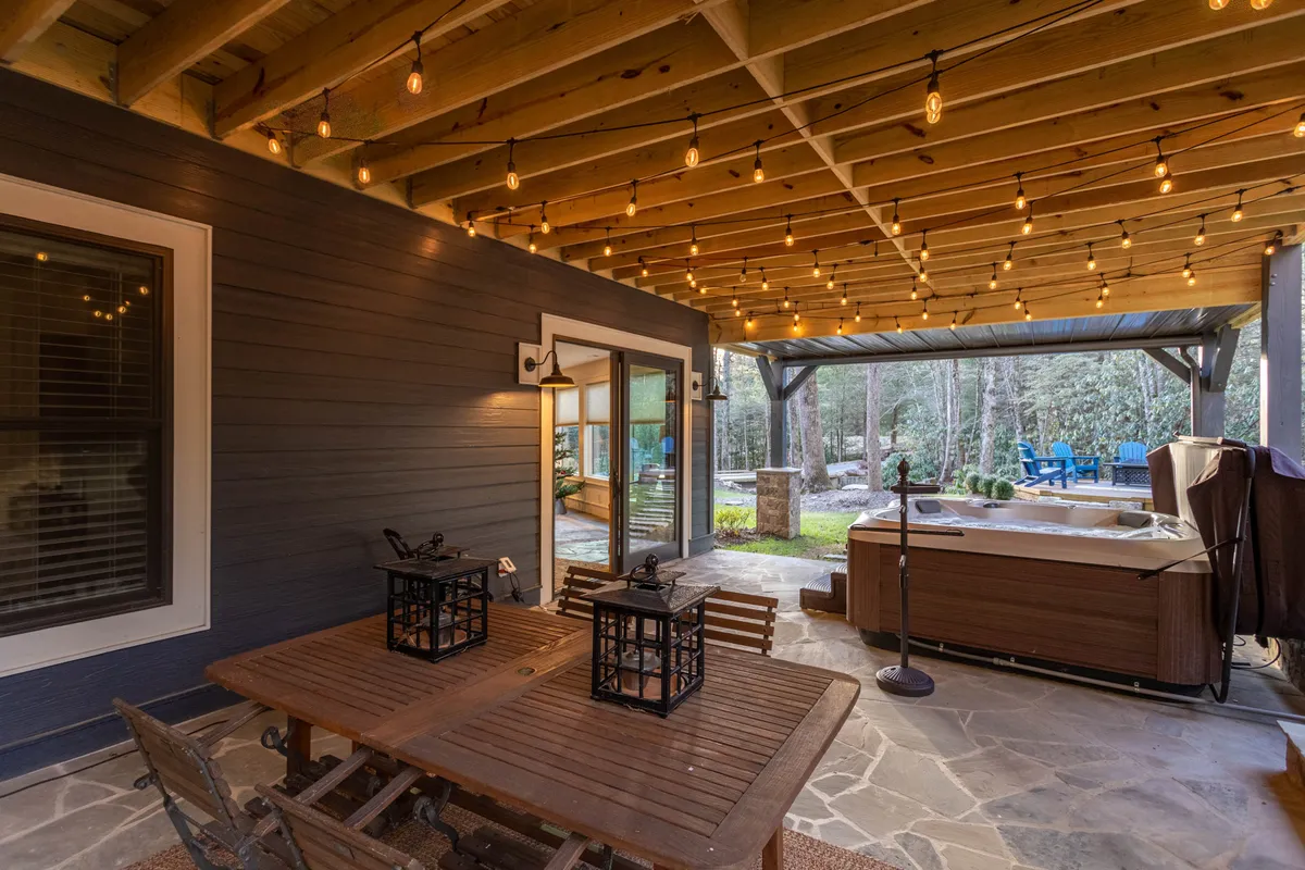 A cozy outdoor area featuring a wooden dining table, hanging lanterns, hot tub, and inviting seating. The ambiance is relaxed and intimate, ideal for unwinding after mountain adventures or gathering under the stars.