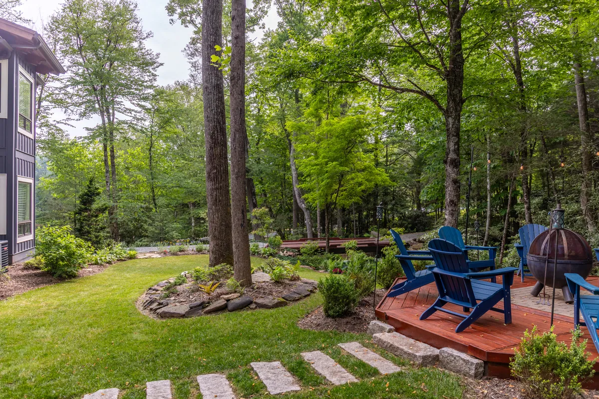 A cozy deck setting with comfortable seating, a fire pit surrounded by lush greenery, and a stone pathway leading into the landscape. This space is clearly designed for gathering, storytelling, and enjoying the natural mountain environment.