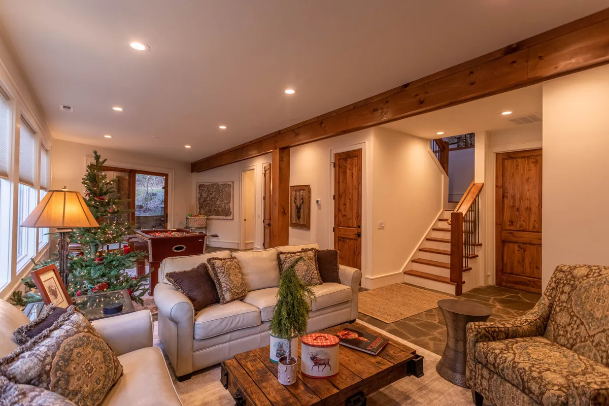 A warmly lit living room featuring a comfortable sofa, a rustic coffee table, a decorated Christmas tree, and a staircase in the background. The space is arranged for gathering, relaxation, and storytelling by the fireplace, evoking a sense of mountain coziness and togetherness.