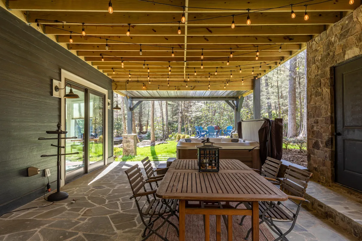 This image features an inviting outdoor dining table under string lights, a hot tub, and a wooden beam structure, all set in an open grassy area. The scene evokes a sense of togetherness, relaxation, and celebration in a mountain setting, perfect for group gatherings.