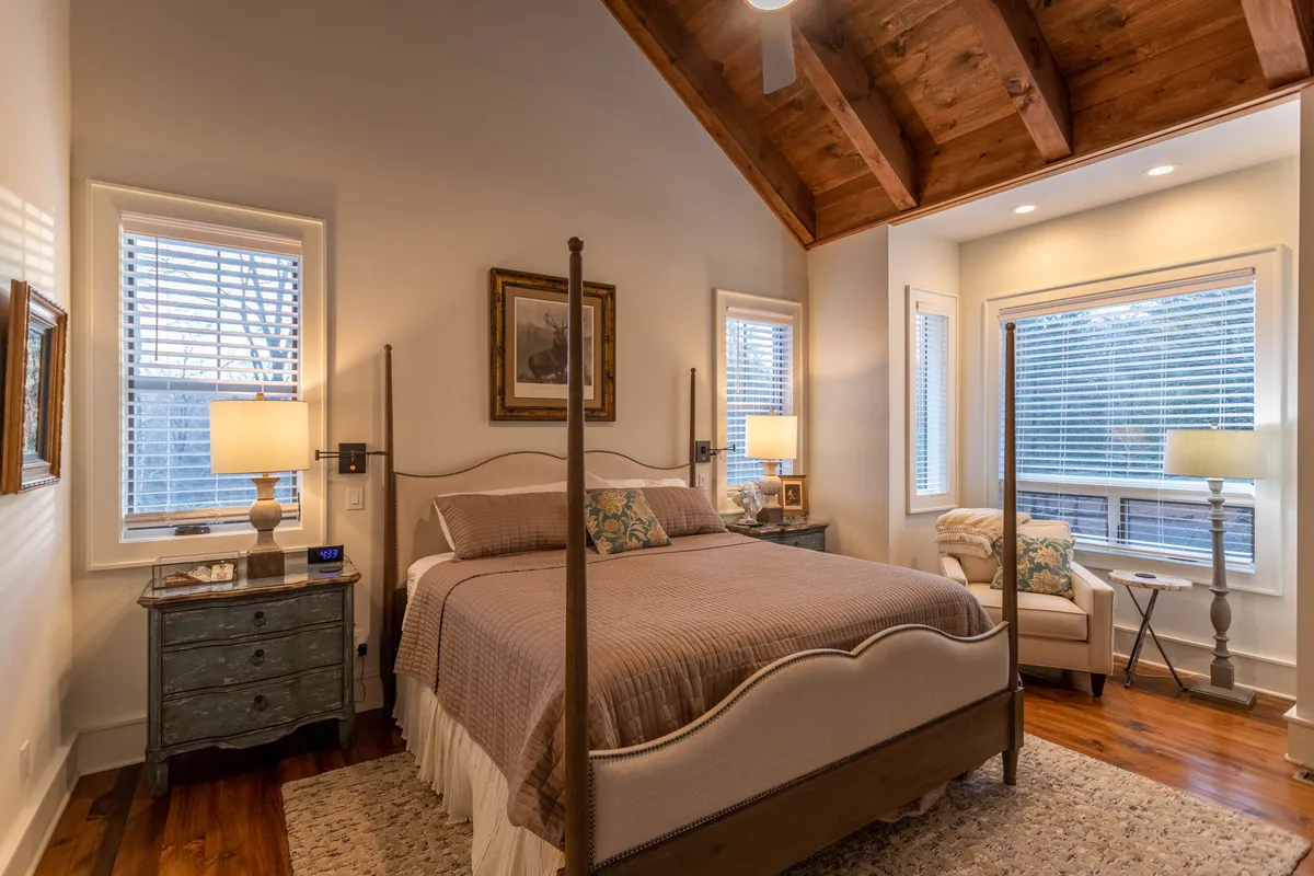 A tranquil king bedroom with a four-poster bed, cozy armchair, nightstand with lamp, and framed artwork. The room is bathed in natural light, creating a peaceful atmosphere for reading or unwinding.