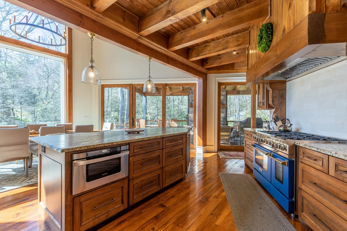 A close-up view of the kitchen area, centering on the grand island, a professional-grade 6-burner gas range, and generous granite countertops. Large windows highlight the natural surroundings and fill the space with light, while the layout invites guests to gather and participate in meal preparation.