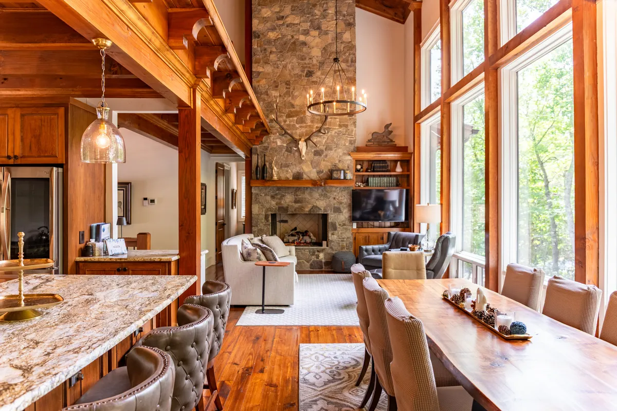 A spacious great room featuring a dramatic stone fireplace, floor-to-ceiling windows flooding the area with natural light, a large dining table set for a group, and an adjacent kitchen island. Plush seating is arranged for conversation and relaxation, and the open-plan layout creates a welcoming flow between the living, dining, and kitchen areas.