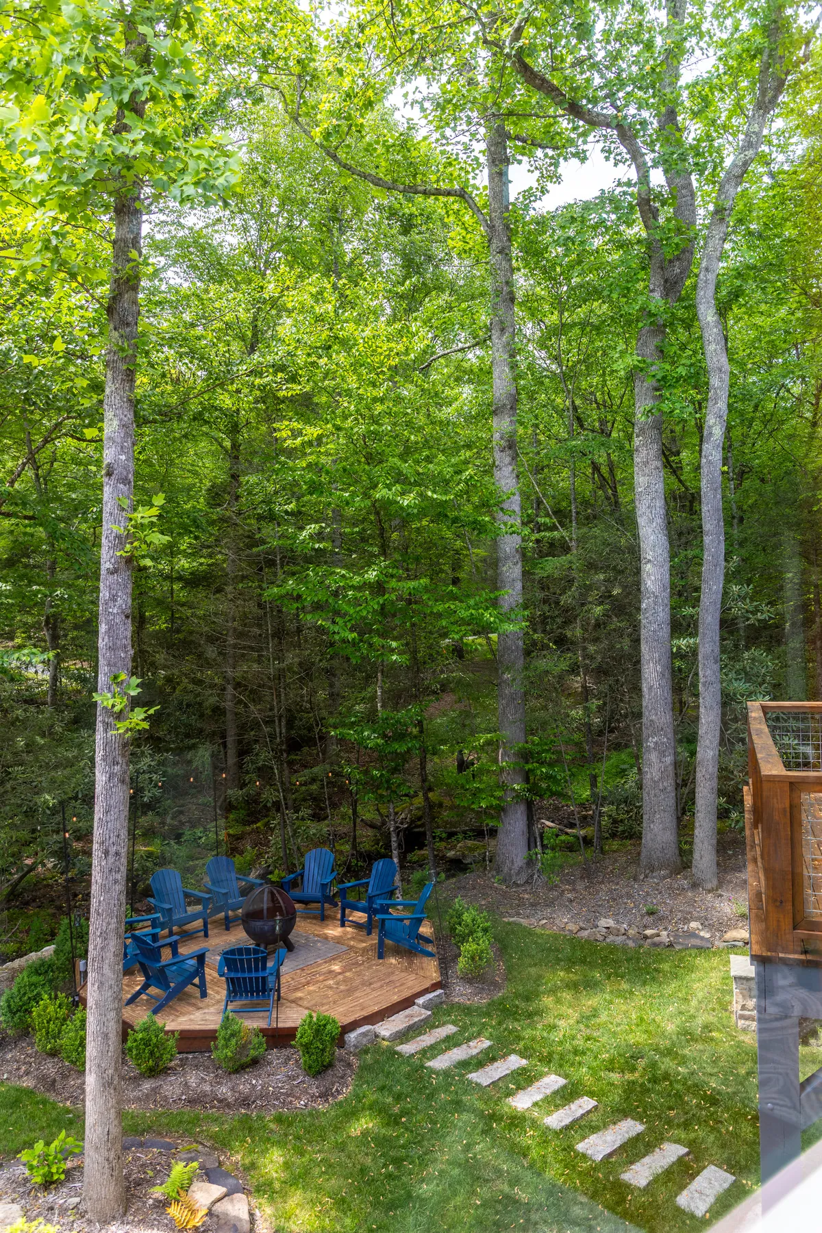 An exterior fire pit area surrounded by blue Adirondack chairs, wooden decking, and a stone pathway. The setting is perfect for evening gatherings, with nature all around and space for friends and family to share stories by the fire.