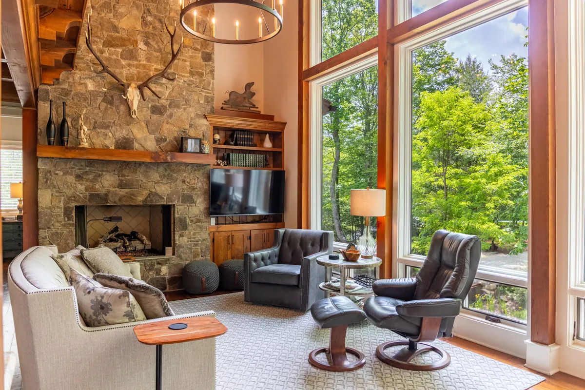 A comfortable seating area set beside a substantial stone fireplace, with large windows letting in dappled sunlight and offering tranquil views of the woods. The decor is warm and inviting, with hints of rustic elegance and a focus on comfort.