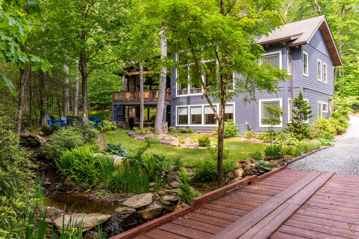A picturesque view of the house set amongst trees and a landscaped garden, with a wooden bridge and a stream in the foreground. The scene is peaceful and inviting, ideal for enjoying outdoor meals, relaxing by the water, or gathering with friends on the deck.