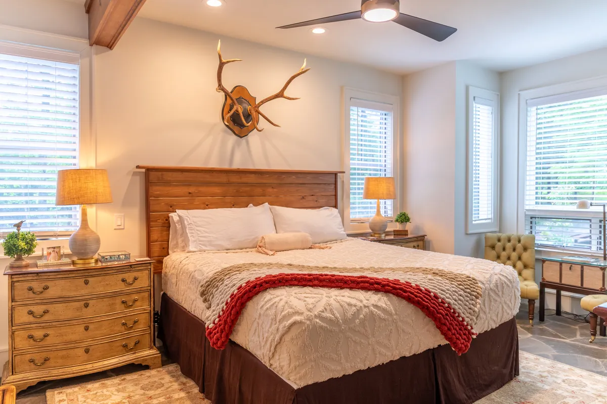 A warm and welcoming king-sized bedroom featuring a wooden headboard with antler accents, decorative lamps, a plush throw blanket, and a sitting area with a comfortable chair. Natural light streams in, creating an inviting atmosphere for rest and relaxation.