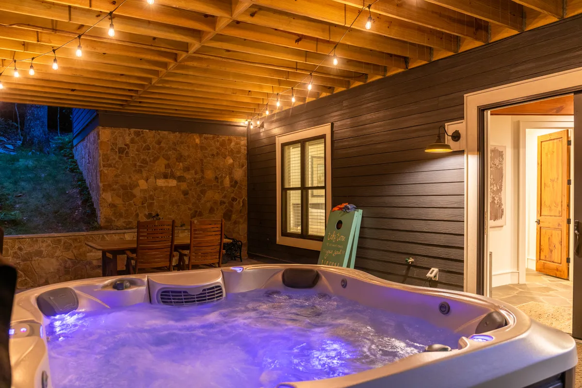 A cozy, indoor hot tub is illuminated by soft lighting and complemented by string lights overhead and a wooden dining set nearby. The mood is warm and relaxing, inviting guests to unwind after adventurous days.