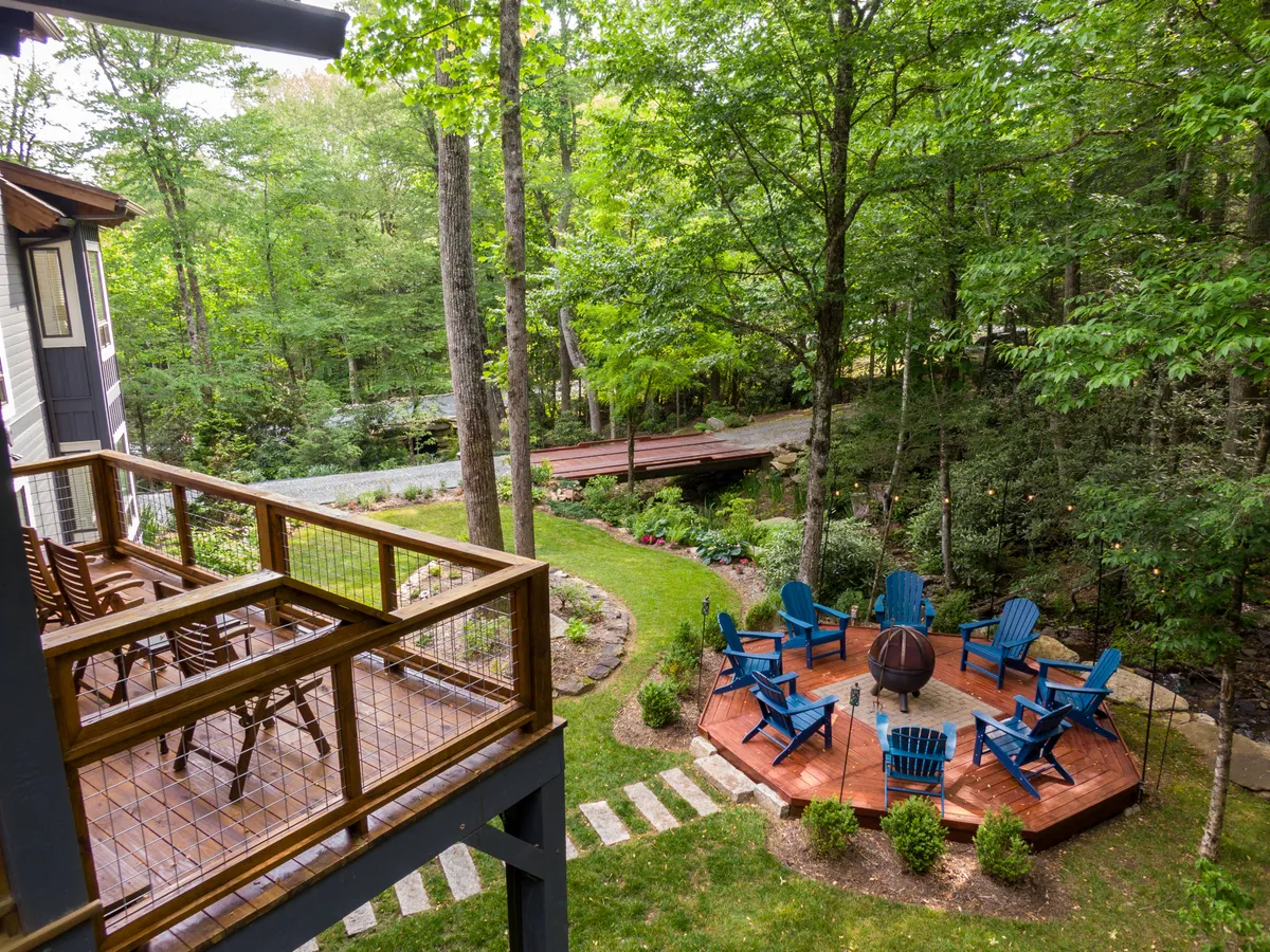 A spacious balcony with comfortable seating overlooks a circular fire pit area, surrounded by greenery and adjacent to a rustic wooden bridge. The scene is perfect for family barbecues, group gatherings, and conversations around the fire in a serene natural setting.