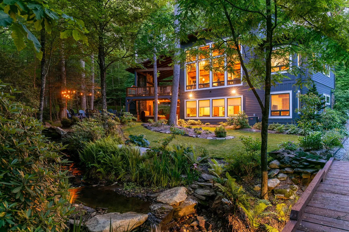 The hero image showcases a beautiful modern home with expansive windows set amidst lush greenery. A picturesque garden and a tranquil creek run beside the house, with soft outdoor lighting creating a warm, inviting glow. The spacious porch invites guests to relax and soak in the peaceful mountain surroundings.
