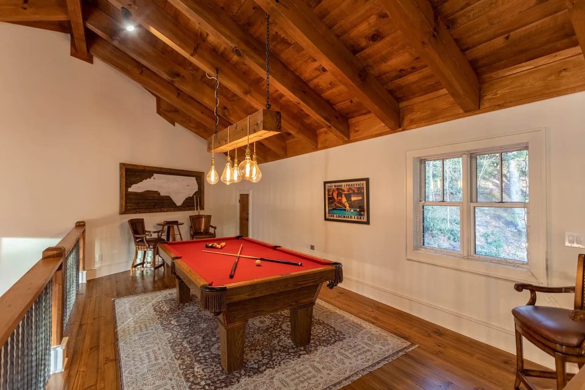 This image features a stylish game loft with a pool table as the focal point, framed by wooden beams, a chandelier overhead, and windows allowing natural light. The atmosphere is casual and lively—perfect for friendly competition and socializing.