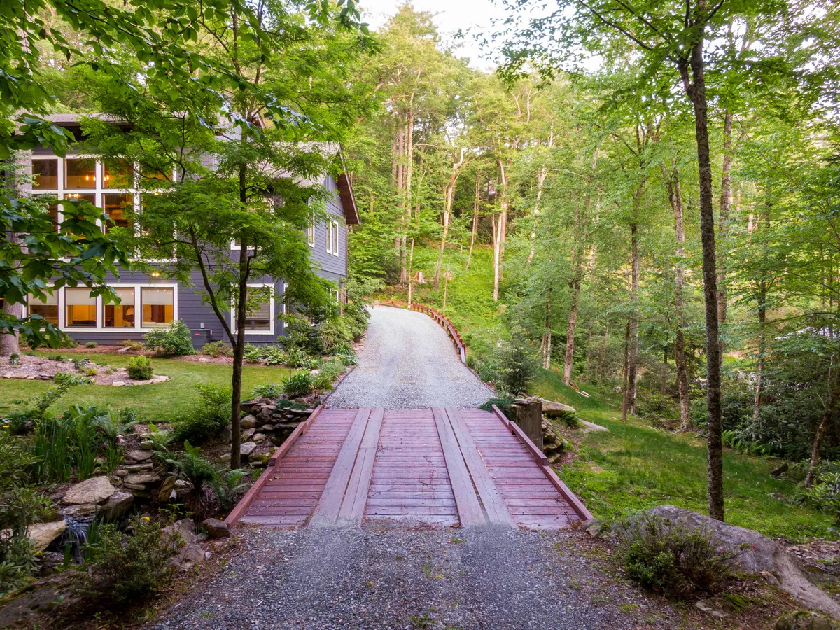 This image displays the home's exterior surrounded by lush greenery, featuring a wooden bridge and a gravel driveway. The scene is serene and welcoming, with clear opportunities for walking through the garden, enjoying nature, and exploring local trails right from the property.