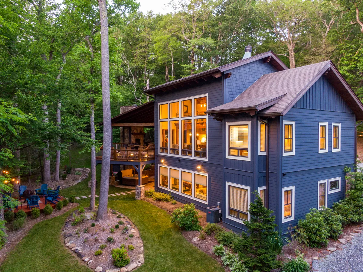 This image features the home's large windows and a wooden deck nestled among lush greenery and manicured gardens. The inviting deck space is ideal for relaxing or gathering, and the expansive windows connect the interior with the tranquil mountain surroundings.