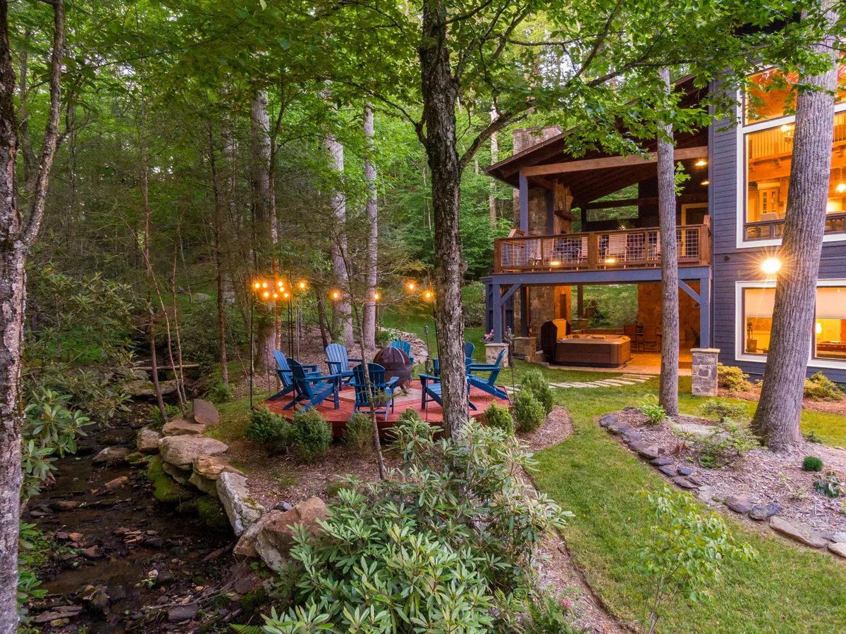 A welcoming wooden deck with contemporary seating, strung with warm string lights, is set beside a gentle stream and surrounded by lush green foliage. The adjacent creek and natural environment invite outdoor relaxation and family picnics, while the comfortable setup hints at al fresco meals and stargazing.